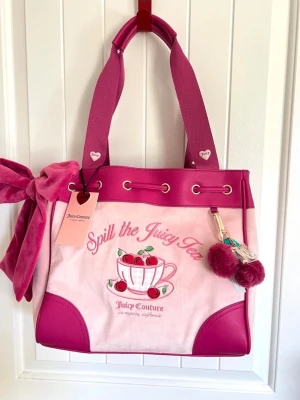 Juicy Couture daydreamer rosa tote - Superfin rosa handväska från Juicy Couture med broderat körsbärsmotiv och texten 'Spill the Juicy Tea'. Väskan har breda axelband, dekorativ rosett i sammet och en söt pompom-nyckelring. Materialmix av tyg och konstläder i olika rosa nyanser.