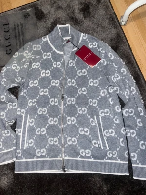 Gucci cardigan  - Snygg grå ziptröja från Gucci med vitt GG-monogram över hela tröjan. Tröjan har ståkrage, vita detaljer vid muddar och fickor samt dragkedja framtill. Perfekt för dig som gillar exklusiv streetstyle och vill sticka ut med lyxigt märke.