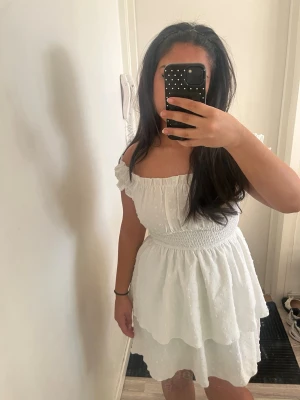 Vit offshoulder kortklänning - Söt vit kortklänning med offshoulder och volangdetaljer. Klänningen har smockad midja och dubbla lager i kjolen för extra volym. Perfekt för varma dagar och har en lätt, luftig känsla.