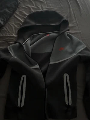 Nike tech kofta - Snygg grå hoodie från Nike med dragkedja framtill och två fickor med vita detaljer. Hoodien har en stor huva och en röd Nike-logga på bröstet. Tillverkad i mjukt material som passar perfekt till en avslappnad stil.