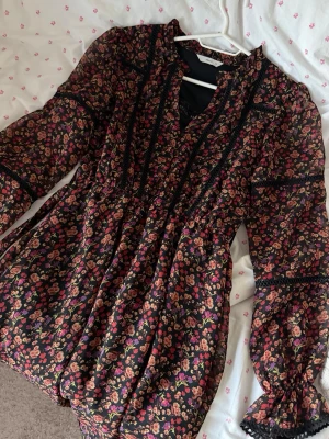Blommig svart klänning med spetsdetaljer -  kortklänning med svart botten och små blommor i rött, rosa och lila. Klänningen har långa ärmar med spetsdetaljer och resår vid ärmslut. V-ringad hals och dekorativa spetsband framtill.