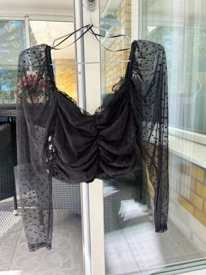 Svart meshblus med puffärmar - Trendig svart blus i mesh med långa puffärmar och prickigt mönster. Toppen har rynkade detaljer framtill och en croppad passform. Perfekt för dig som vill sticka ut med en cool och stilren look.