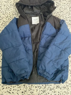 Blå och svart pufferjacka Jack & Jones - Snygg blå och svart pufferjacka från Jack & Jones med huva och dragkedja. Jackan har breda kanaler, elastiska muddar vid ärmslut och är fodrad för extra värme. Perfekt för kalla dagar och enkel att matcha med olika outfits.