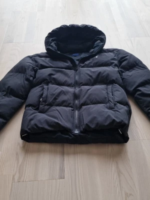 Svart pufferjacka från GANT - Snygg svart pufferjacka från GANT med huva och dragkedja framtill. Jackan har två stora fickor med lock och är vadderad för extra värme. Perfekt för kalla dagar och har en klassisk, stilren look. Storlek 176 men sitter som en small för man!