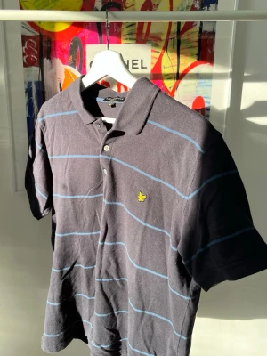 Svart randig pikétröja Lyle & Scott - Klassisk svart pikétröja från Lyle & Scott med tunna blå ränder och gul logga på bröstet. Tröjan har krage, knappar framtill och korta ärmar. Perfekt för en avslappnad och stilren look. Storlek XL i junior dvs M i senior. 