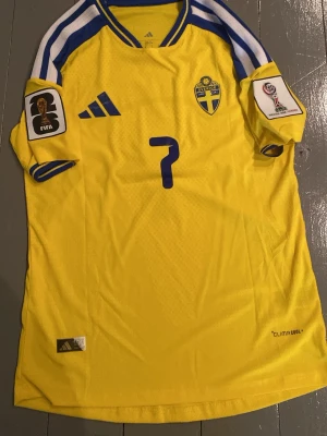 Svensk landslagströja Adidas nr 7 - Gul svensk landslagströja från Adidas med blå detaljer och nummer 7. Tröjan har officiella FIFA och turneringsmärken på ärmarna, samt 'Bergvall' tryckt på ryggen. Tillverkad i lätt och ventilerande ClimaCool-material, perfekt för fotboll.