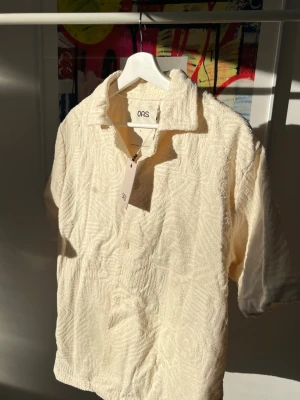 Beige frottéskjorta från OAS - Säljer en beige skjorta från OAS i mjuk frotté med korta ärmar och öppen krage. Skjortan har ett subtilt mönster i tyget och en avslappnad passform, perfekt för varma dagar. Märkeslapp och tagg kvar. Den är lite stor i storleken och kan upplevas något ’oversized’.