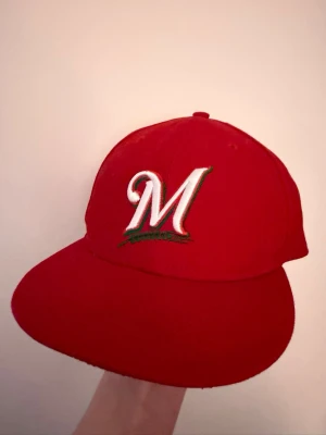 Röd keps med broderat M - Säljer en röd keps med platt skärm och ett broderat vitt M med grön och svart detalj framtill. Klassisk snapback-modell som ger en sportig vibe. Perfekt för dig som vill sticka ut med en färgstark accessoar.