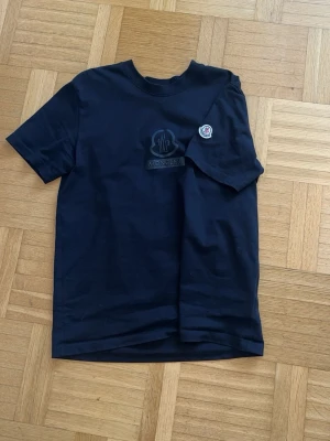 Svart Moncler t-shirt med logga - Snygg svart t-shirt från Moncler med klassisk rund hals och korta ärmar. T-shirten har en diskret Moncler-logga broderad på bröstet och en patch på ärmen. Tillverkad i mjuk bomull för skön känsla och stilren look.