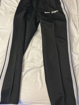 Palm Angels svarta mjukisbyxor - Svarta mjukisbyxor från Palm Angels med vita sidoränder och logga framtill. Byxorna har rak passform och är tillverkade i ett mjukt, stretchigt material som ger en sportig vibe. Perfekta för en avslappnad och trendig look. Priset är diskuterbart