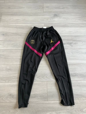 Svarta PSG x Jordan träningsbyxor - Säljer ett par svarta träningsbyxor från Jordan x PSG med guldiga loggor och lila detaljer över låren. Byxorna har elastisk midja och smal passform, perfekta för fotboll eller gym. Materialet är troligtvis polyester för maximal rörelsefrihet.