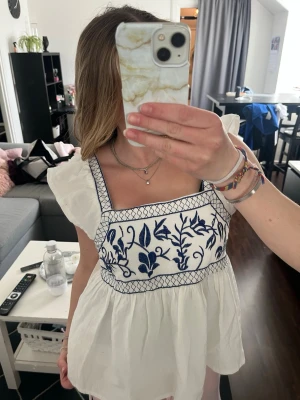 Vit blus med blå broderi och volang - Supersöt vit blus med blå broderade blommor och volangärmar. Fyrkantig ringning och breda axelband med broderade detaljer och knytning bak. Luftig och flowy passform, perfekt för varma dagar. Använd 1 gång. 