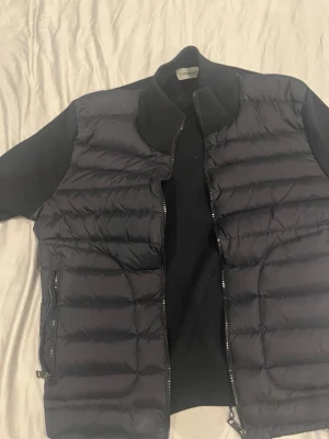 Svart dunväst från Moncler - Snygg svart dunväst från Moncler med quiltad framsida och dragkedja. Västen har ribbade detaljer vid kragen och två dragkedjefickor. Klassisk Moncler-logga på ärmen och stilren design som passar perfekt till lager-på-lager-look.
