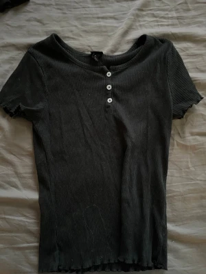 Svart ribbad topp med knappar - Svart ribbad topp med korta ärmar och tre vita knappar framtill. Toppens material är mjukt och stretchigt, och den har en rund halsringning. Perfekt för en chill och stilren look.