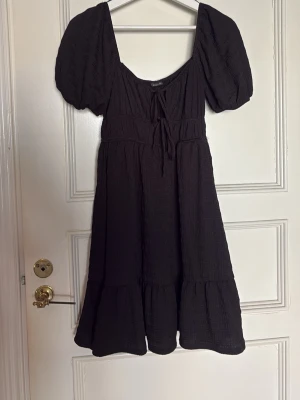 Svart kortklänning från Gina Tricot - Supersöt svart klänning från Gina Tricot med puffärmar och knytning framtill. Klänningen har en volang nertill och är tillverkad i ett strukturerat tyg som ger en trendig look. Endast använd 1 gång 💕