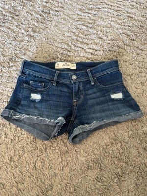 Mörkblå jeansshorts - Hollister - Snygga mörkblå jeansshorts från Hollister med slitna detaljer och broderi på bakfickorna. Modell med låg midja och knappgylf. Perfekta för sommaren! ☀️strl. 00/w23, kolla bilder m. mått. Säljes då dem tyvärr börjar bli försmå för mig. Pris går att diskutera, fraktas. Säljes i befintligt skick. Köpta ca 2024.