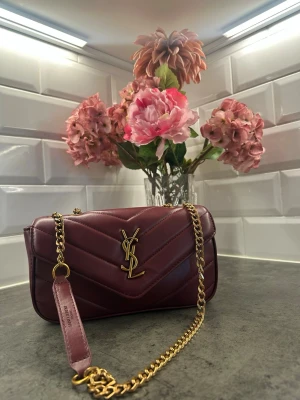 Vinröd YSL handväska i läder - Snygg vinröd handväska från YSL i quiltat läder med guldiga detaljer och kedjerem. Väskan har klassisk YSL-logga framtill och en stilren, rektangulär form. Perfekt accessoar för att lyfta din outfit.