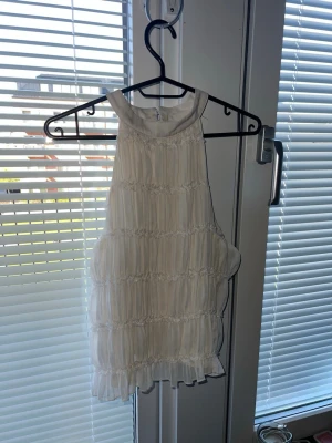 Vit ärmlös blus med volangdetaljer - Säljer en söt vit blus med hög hals och volangdetaljer. Toppen är ärmlös och har en luftig, lätt struktur med fina rynkade partier. Perfekt för dig som gillar en romantisk och fräsch stil.