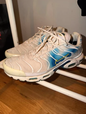 Nike TN  - Nike Air Max Plus sneakers i vitt och blått med gula detaljer. Skorna har det klassiska TN-mönstret och synlig Air-sula. Materialet är en mix av mesh och syntet, med vita snören och en gul Swoosh på sidan. Perfekta för dig som gillar retrostil och streetwear.