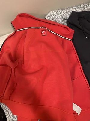 Röd hoodie från Nike med huva - Snygg röd hoodie från Nike med klassisk huva och vit detalj längs axlarna. Hoodien har en sportig look och är tillverkad i mjukt material, perfekt för en avslappnad stil. Ikonisk Nike-logga på insidan av kragen.