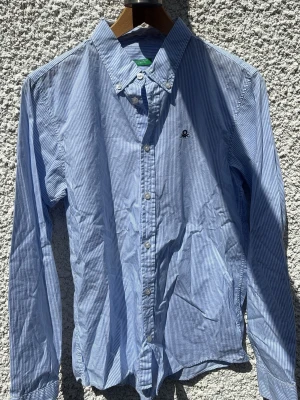 Blårandig skjorta från Benetton - Klassisk blårandig skjorta från United Colors of Benetton med button down-krage och broderad logga på bröstet. Skjortan har långa ärmar och stängs med vita knappar framtill. Perfekt för en stilren look.