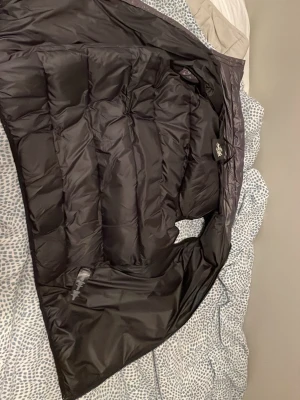 Svart dunväst från The North Face - Snygg svart dunväst från The North Face med quiltad design och hög krage. Västen har en glansig yta upptill och matt tyg nedtill, perfekt för lager-på-lager. Klassisk logga på insidan och dragkedja i sidan.