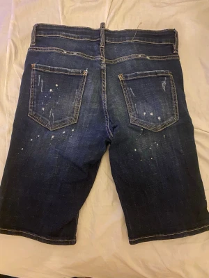 Mörkblå jeansshorts från Dsquared2 - Snygga mörkblå jeansshorts från Dsquared2 med slitna detaljer, vita färgstänk och klassisk femficksdesign. Shortsen har normal passform och är tillverkade i bomull med en cool streetkänsla. Perfekta för dig som gillar en trendig och avslappnad stil.