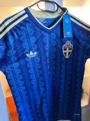 Blå Sverige tröja från Adidas - Blå matchtröja från Adidas med Sveriges landslagsemblem på bröstet och klassiska Adidas-ränder i gult på axlarna. Tröjan har ett mönstrat tyg och V-ringning, tillverkad i polyester. Perfekt för fotboll eller träning.