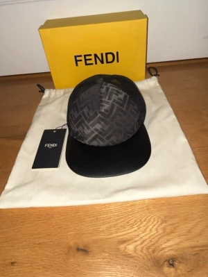 Svart keps från Fendi med FF-mönster - Helt ny Snygg svart keps från Fendi med klassiskt FF-mönster på framsidan och mesh-tyg baktill. Justerbar rem med guldfärgad Fendi-logga bak. Skärmen är i svart mockaimitation och kepsen har en modern, sportig look.