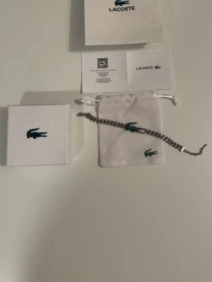 Lacoste silverfärgat armband - Snyggt armband från Lacoste i silverfärgad metall med en ikonisk grön krokodil i mitten. Krokodilen är dekorerad med små pärlor i grönt och vitt samt en röd detalj vid munnen. Kommer med original ask och tygpåse.