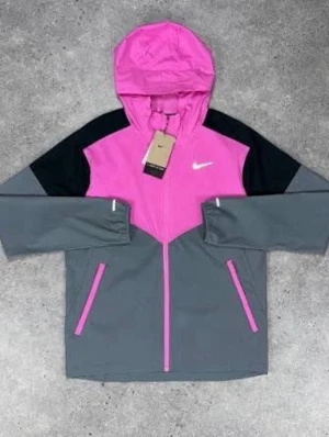 Nike vindjacka rosa och grå - Snygg vindjacka från Nike i en kombination av rosa, grått och svart. Jackan har huva, dragkedja framtill och två fickor med rosa detaljer. Perfekt för dig som vill ha en sportig och färgstark look. Nike-logga på bröstet och modern passform.