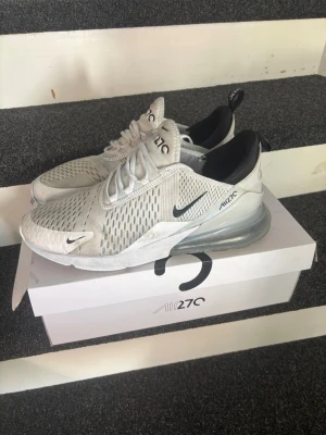 Nike Air Max 270 vita sneakers - Nike Air Max 270 sneakers i vitt med svarta detaljer och stor synlig Air-enhet i hälen. Ovandelen är i mesh för bra ventilation och har en sportig, modern look. Klassisk Nike-logga på sidan och tungan. Perfekta för dig som gillar streetstyle och bekväma skor.