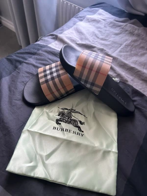 Burberry slides med klassiskt rutmönster - Säljer ett par Burberry slides med svart sula och bred rem i det ikoniska beige, svarta och röda rutmönstret. Tillverkade i syntetmaterial med bekväm fotbädd. Perfekta för en trendig och avslappnad stil. Dustbag från Burberry medföljer.