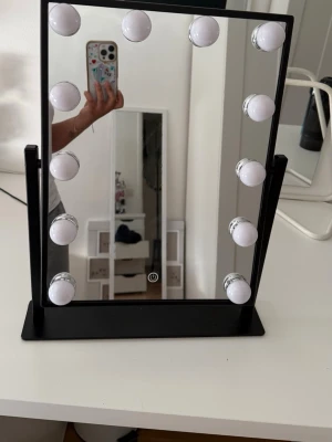 Svart sminkspegel med LED-lampor - Stilren sminkspegel med svart ram och fot, utrustad med tio runda LED-lampor runt spegeln för optimal belysning. Spegeln har en touchknapp på framsidan för att enkelt tända och släcka lamporna. Perfekt för sminkning och styling.