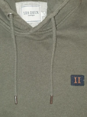 Olivgrön hoodie från Les Deux - Snygg olivgrön hoodie från Les Deux med broderad logga på bröstet och klassisk känguruficka. Hoodien har dragsko i huvan och ribbade muddar vid ärmslut och nederkant. Perfekt för en chill och stilren look.