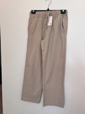 Beige linnebyxor  i strl 160 - Snygga beige linnebyxor från 157 med avslappnad passform och resår i midjan. Byxorna har raka ben och praktiska sidfickor. Perfekta för en chill och stilren look. Materialet är svalt och skönt linne.