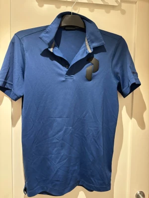 Blå golfpike från Peak Performance | S - Snygg blå pikétröja från Peak Performance med klassisk krage och korta ärmar. Tröjan har diskret logga broderad på ärmen och är tillverkad i ett mjukt och stretchigt material som känns bekvämt mot huden.