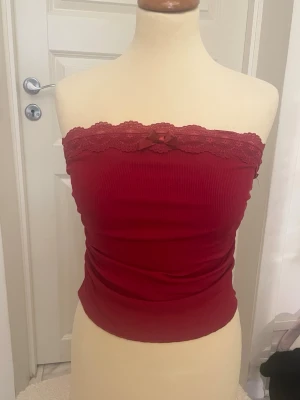 Röd bandeau tubtopp med spets - Snygg röd bandeau tubtopp med ribbad struktur och spetskant upptill. Toppen har en liten rosett framtill och är axelbandslös, perfekt för en trendig look. Materialet känns mjukt och stretchigt för en bekväm passform. Passar S oxå.