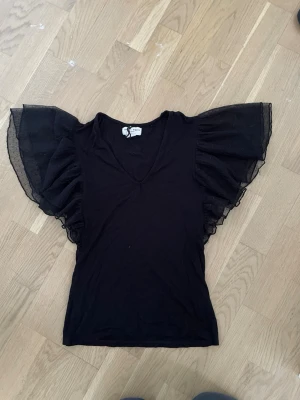 Svart topp med volangärmar Holly & Whyte - Snygg svart topp från Holly & Whyte by Lindex med djup V-ringning och stora volangärmar i transparent mesh. Perfekt för dig som vill ha en trendig och unik look. Materialet känns mjukt och lätt, och ärmarna ger toppen en extra cool touch.