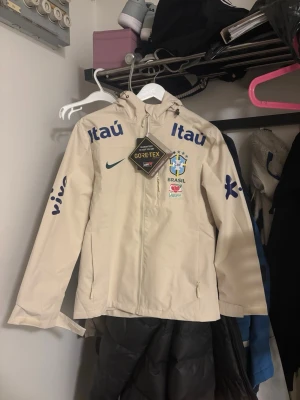 Beige Brasil Nike vindjacka GORE-TEX - Stilren beige vindjacka från Nike med Brasil-logga, GORE-TEX-material och tryckta sponsorloggor. Jackan har huva, hel dragkedja och fickor med dragkedja. Perfekt för dig som gillar sportig stil och vill sticka ut.