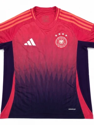 Tysklands fotbollströja Adidas röd/lila - Snygg Tyskland fotbollströja från Adidas i röd och lila med gradient mönster. Tröjan har rund hals, korta ärmar och tre ränder på axlarna. Tysklands landslagsmärke och fyra stjärnor på bröstet. Tillverkad i lätt och ventilerande AEROREADY-material.