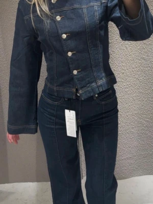 Jeans set - Trendig mörkblå jeansjacka oc med asymmetrisk knäppning och raka, vida ärmar. Jackan har rund halsringning och tydliga kontrastsömmar. Jeansen är i storlek 34 och tröjan xs. Perfekt för dig som vill sticka ut med en unik denimlook. Endast använd en gång!
