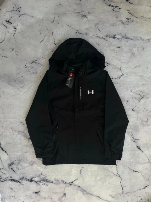Svart vindjacka från Under Armour - Svart vindjacka från Under Armour med huva och dragkedja framtill. Jackan har två sidofickor med dragkedja, elastiska muddar och en vit logga på bröstet. Materialet är lätt och vattenavvisande, perfekt för blåsiga dagar.