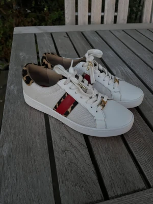 Vita sneakers med leoparddetaljer - Snygga vita sneakers med röda och leopardmönstrade ränder på sidorna. Skorna har vita snören, guldfärgade detaljer och leopardmönstrad häl. Perfekta för sommaren och våren!