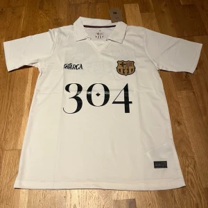 Barcelona X lamine yamal special edition  - Snygg vit kortärmad tröja från FC Barcelona med svart tryck '304' på framsidan och klubbmärke i guld. På ryggen finns ett stort tryck med två porträtt och texten 'Lamine Yamal'. Perfekt för dig som älskar fotboll och vill sticka ut.