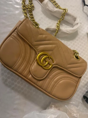 Beige  handväska - Snygg beige Marmont handväska i quiltat läder med guldfärgad kedja och GG-logo framtill. Väskan har ett innerfoder i vinrött tyg och dragkedjefack. Perfekt för dig som vill ha en trendig och lyxig accessoar.