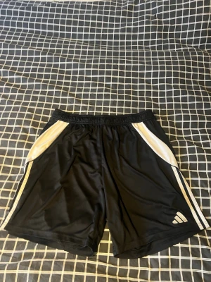 Svarta Adidas shorts med vita ränder - Svarta sportiga shorts från Adidas med vita ränder längs sidorna och vit detalj vid fickorna. Klassisk Adidas-logga på vänster ben. Perfekta för träning eller chill. 