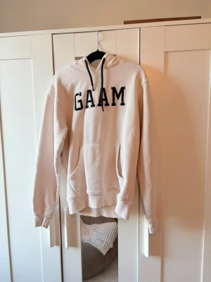 Beige hoodie från GAAM - Snygg beige hoodie från GAAM med svart broderad logga på bröstet. Klassisk modell med huva, känguruficka och ribbade muddar. Perfekt för en chill och avslappnad stil. Mjuk bomullsblandning som känns skön mot huden.