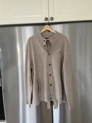 Beige kofta med knappar och fickor - Mysig beige kofta med ribbad krage och knappstängning framtill. Två fickor på sidorna och diskret logga på bröstet. Långärmad modell i mjukt stickat material, perfekt för lager på lager och chill stil.