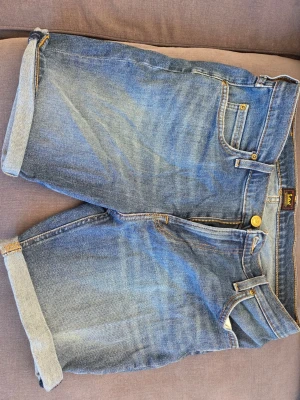 Blå jeansshorts från Lee - Snygga blå jeansshorts från Lee med klassisk femficksdesign och uppvikta benslut. Shortsen har gylf med dragkedja och knapp, samt tydliga kontrastsömmar. Perfekta för varma dagar och enkel att matcha med t-shirt eller hoodie.